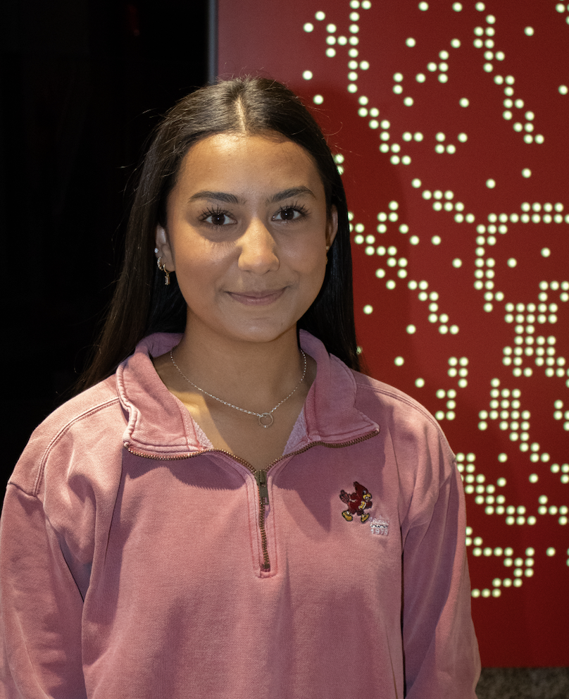 Radhika Sharma, co-director of the 2024 ISU Homecoming Central Committee, portrait in front of red wall with circle lights.